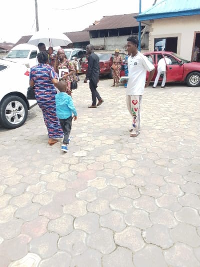 The image shows a group of people standing in a paved area in Yenagoa, Nigeria, located at coordinates 4.931 latitude and 6.267 longitude. There are six individuals visible in the photo: a woman in a blue and white patterned dress holding a white bag, a boy wearing a blue shirt and jeans, a woman holding a brown bag, a man in a black shirt and pants, and a man wearing a white shirt with colorful pants. The man in the white shirt is holding a white book with a picture of a heart on the cover. There are a couple of parked vehicles in the background, one of which is red and has a flat tire on the back left wheel. The scene is a paved area with a cloudy sky and a few buildings visible in the background. The paved area appears to be in front of an apartment complex.