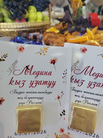 A close-up shot captures two gold-wrapped chocolates placed on white cards adorned with floral designs and text in a cursive script. The left card features the text "Медина кыз узатуу" and below it, "Биздин жакшылыдыгыды тен болушконунуз учун Рахмат!" followed by a date. The right card appears to have similar text. The chocolates are positioned at the bottom of each card. In the background, out of focus, a platter of fruits, including grapes and apples, is visible, as well as other food items, suggesting a celebratory event or gathering. The scene likely takes place in a domestic setting within Бактуу-Долоноту, Kyrgyzstan, given the provided context.