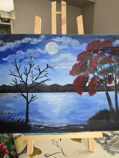 This is a painting of a moonlit landscape. The sky is a deep blue with fluffy white clouds and a full moon that casts a yellowish glow. In the foreground, a calm body of water reflects the moonlight. On the left side of the water, there is a bare, dark tree with a bird perched on one of its branches. In the background, silhouetted mountains rise against the sky. On the right side of the water, a tree with vibrant red leaves is depicted. The painting is displayed on a wooden easel. The background behind the easel appears to be a wall and a shelf with some objects on it, possibly in a room in Claremorris, Ireland.