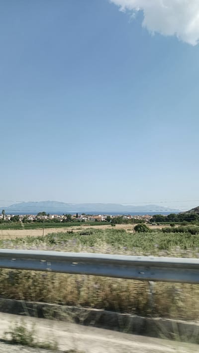 The image captures a distant view of Nea Karvali, Greece, seen across a body of water and coastal landscape. In the foreground, a blurred metal guardrail runs horizontally across the lower portion of the frame, indicating the photo was taken from a moving vehicle. Beyond the guardrail, a field of dry grass and scattered green vegetation stretches towards a more cultivated area with rows of crops. A town with numerous houses featuring red-tiled roofs is visible along the coastline. Behind the town, a long, low mountain range is silhouetted against the clear blue sky. A single white cloud is present in the upper right corner. The scene is bathed in bright daylight, suggesting a sunny day.