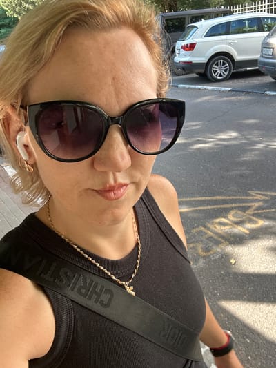 A woman with blonde hair and sunglasses is taking a selfie outdoors. She is wearing a black tank top and a gold necklace. A white wireless earbud is visible in her left ear. In the background, there are cars parked on a street. The image appears to be taken during the daytime, with sunlight casting shadows on the asphalt. The location is identified as Aderbiyevka, Russia.