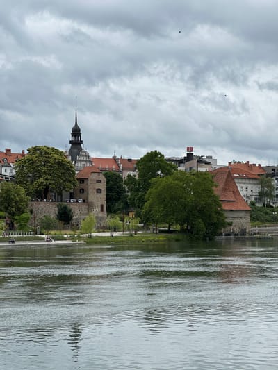 The image depicts a waterfront view of Maribor, Slovenia, focusing on the Old Vine House and the Maribor Castle Tower. The scene is captured on an overcast day, with a cloudy sky dominating the upper portion of the frame.
In the foreground, a wide river, likely the Drava River, flows with gentle ripples, reflecting the muted light. The water's surface shows texture and depth.
The middle ground reveals a grassy embankment lined with trees and a paved walkway. Several people are visible on the embankment. Two individuals are seated on a bench near the water, while a person on a bicycle is further along the path. In the background, the architectural highlights of Maribor are prominent.
To the left, the stone walls of a historic building, identified as the Maribor Castle Tower (part of the Maribor Castle complex), stand with arched windows. Adjacent to it, a large, mature tree with lush green foliage provides shade. Beyond this, a cluster of buildings with red-tiled roofs and the distinct spire of a church tower are visible, indicating the city center.
To the right, another section of a stone building with a prominent red-tiled roof, likely part of the Old Vine House complex, is featured. Further back, modern buildings, including one with a red logo, punctuate the skyline. The overall impression is of a historic European city by a river, under a typically cloudy sky.