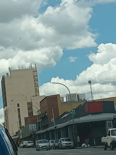 The image depicts a daytime street scene in an urban commercial area of Germiston, South Africa. The sky is partially cloudy with distinct white cumulus clouds against a blue background.
In the background and midground, a row of multi-story buildings lines the street. On the left, a tall, light-colored building with a flat roof features several communication antennas. Adjacent to it and extending to the right, a series of commercial structures are visible. These buildings vary in facade, including some with red brick and others with darker finishes, many equipped with corrugated metal awnings extending over what appear to be storefronts. One building on the right exhibits a prominent horizontal red band near its roofline.
The foreground and midground show a paved road occupied by multiple vehicles. A line of cars, predominantly light-colored sedans and possibly SUVs or light trucks, are either stopped in traffic or parked along the street. A white pickup truck is visible on the far right. A blue vehicle partially obstructs the lower left portion of the image, suggesting the photograph was taken from within a vehicle.
Street infrastructure includes a tall, curved streetlight extending over the road and another pole with a spherical light fixture on the right. A green street sign displaying the number "5" is mounted above the commercial buildings. No individuals are clearly discernible within the frame.