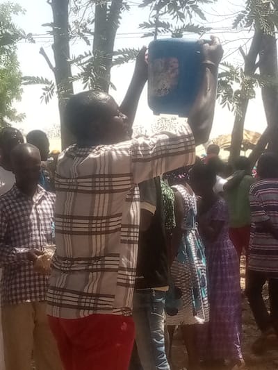 In Kagoma, Nigeria, an outdoor scene unfolds under bright daylight. A person with dark skin, wearing a light-colored plaid long-sleeved shirt and red trousers, stands prominently in the foreground, facing right but looking upwards. This individual holds a blue plastic jerrycan, approximately 5-10 liters in capacity, above their head with both hands, appearing to either pour liquid or access its contents. The top of the person's head and parts of their upper body appear wet, consistent with water or another liquid. In the blurred background, a crowd of approximately 10-15 individuals, including adults and children dressed in various casual attire, such as plaid shirts and patterned dresses, are visible. The environment features sparse trees with light green foliage and an unpaved earthen ground. A light-colored, possibly temporary, structure or tent is discernible in the far right background.
