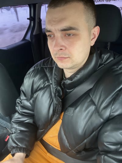 A male individual is seated in the front passenger seat of a vehicle in Novosinkovo, Russia. He has short, dark hair, light facial stubble, and light brown eyes, with his gaze directed downwards and to the right. He wears a black, quilted, synthetic leather puffer jacket, partially zipped, over an orange garment. A black seatbelt is buckled across his chest. The vehicle's interior is dark, with a patterned fabric visible on the seat to the subject's left. Through the blurred side window, a cold, possibly snowy or overcast outdoor environment is visible, characterized by indistinct white and grey tones and faint linear elements. Partial Cyrillic text, including 'КОНЧД', is faintly discernible on a white background in the upper left of the exterior view.