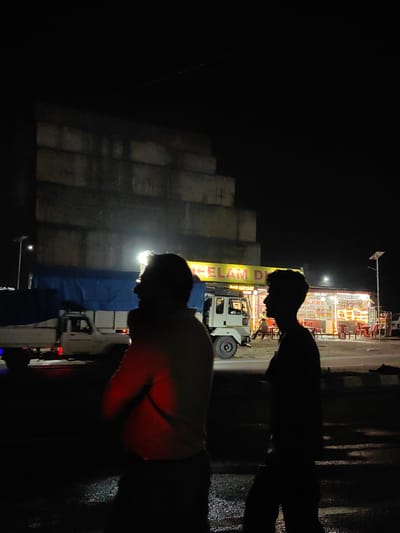The image depicts a nighttime scene on a road in Sambal, India. Two men are silhouetted in the foreground, facing away from the camera. To their right, a brightly lit roadside establishment with tables and chairs is visible, likely a restaurant or dhaba. A truck covered with a blue tarp is parked on the left side of the road, and another vehicle is partially visible further down. The background features a large, dark structure, possibly a building under construction or an unfinished wall. The overall atmosphere is dimly lit, with artificial lights from the establishment illuminating the immediate surroundings.