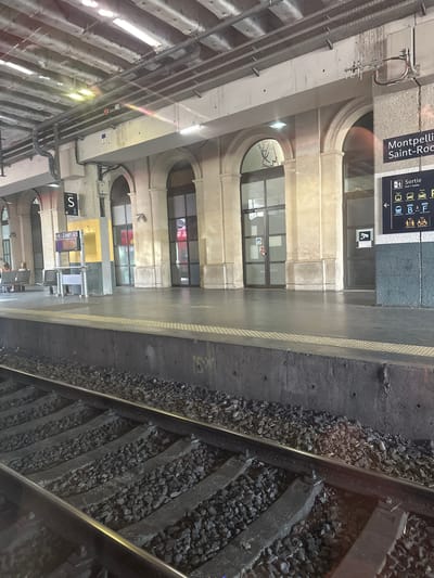 The image shows a train platform at Montpellier Saint-Roch station in Montpellier, France. In the foreground, train tracks with ballast are visible. The platform is made of concrete with tactile paving along the edge. Behind the platform, there are arched doorways and windows, indicative of traditional station architecture. A sign displaying "Montpellier Saint-Roch" is mounted on a dark grey column, with a directional sign below it indicating "Sortie" (Exit) and destinations like "Tram," "Taxis," and "Vélos" (Bikes). Several people are seated on benches on the platform, waiting. The environment appears to be an indoor or covered section of the station, with a visible ceiling structure and lighting. The overall scene captures a moment of waiting at a functional train station.