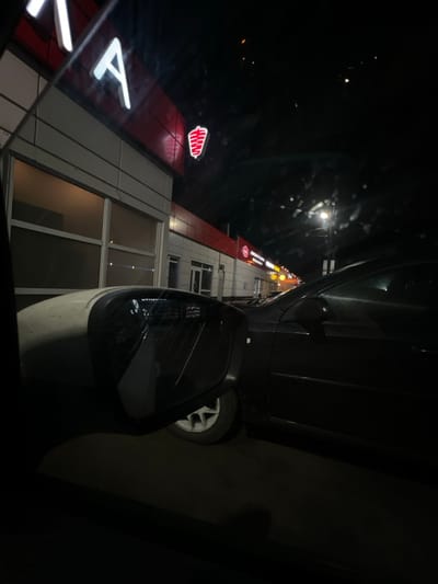 The image is oriented ninety degrees clockwise, depicting a nighttime scene. When viewed upright, the perspective is from inside a vehicle looking out towards a street and a commercial building.

In the immediate foreground, to the bottom right, is the side mirror of a light-colored car, possibly white or light gray. Its reflective surface shows some distortion and light reflections. Part of a dark-colored vehicle, likely the one from which the photo was taken or one parked closely alongside, occupies the bottom left portion of the frame. A wheel with a light-colored rim is visible beneath this dark vehicle.

Beyond the vehicles, a multi-story building with a modern architectural style dominates the midground. Its facade features red and white or light gray paneling, interspersed with large rectangular windows.

Several illuminated signs are visible on the building:
*   On the left side of the building, a large red section displays a prominent white letter "A", though partially cut off by the edge of the frame.
*   Above this "A", there is a smaller, distinct red sign featuring a stylized white logo resembling a 'W' or a battery icon with horizontal lines inside.
*   Further along the building towards the right, a long red banner with white capital letters clearly spells out "ПРОМСВЯЗЬБАНК" (Promsvyazbank), indicating a financial institution.

The overall setting is dark, signifying nighttime. Various bright spots of light are scattered in the background, suggesting streetlights or other illuminated elements further down the road, creating reflections on the car window from which the photo was taken. No specific activity is depicted; it is a static shot capturing the illuminated urban environment at night.