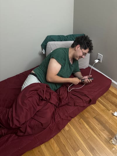 The image shows a young man relaxing indoors, likely in a bedroom or casual living space in Franklin Township, United States of America. He is lying on his left side on a bed, supported by a grey pillow, and partially covered by a dark red comforter or duvet.
The man has curly dark hair and wears glasses. He is dressed in a dark green t-shirt and grey boxer briefs, with the brand "FRUIT OF THE LOOM" visible on the waistband of his underwear. He is actively using a dark-colored smartphone, holding it with both hands and looking at its screen. The phone is connected via a white charging cable to a white charger plugged into an electrical outlet on the wall above his head, suggesting it is being charged while in use.
The room features a two-toned wall, with a darker grey section meeting a lighter grey section lower down. The floor is covered in light-colored wooden planks. Near the edge of the bed on the floor, a crumpled piece of white paper or tissue and a clear bottle of water are visible. The man appears calm and engaged with his device. The indoor setting does not provide specific cues regarding the time of day or weather.