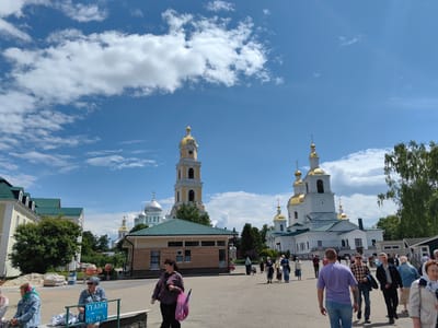This is a wide, eye-level shot of a public square in front of a large religious complex, likely a monastery or cathedral, in Diveyevo, Russia. The architecture features multiple white buildings with green roofs and ornate golden domes, including a prominent bell tower with a clock. A bright blue sky with fluffy white clouds fills the upper portion of the image.
Numerous people are walking in the square, dressed in a variety of casual and modest clothing. Some are walking individually, while others are in small groups. Several women are wearing headscarves. In the foreground, a few people are seated on a bench, one of whom is looking at the camera. A blue sign with white text and icons indicating "Toilet WC" is visible on the bench. There are also stacks of building materials and construction equipment in the middle ground, suggesting ongoing work or renovations. Trees line the edges of the complex and the square, and the overall impression is of a busy, sacred site on a sunny day.