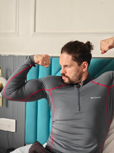 A man with dark hair pulled into a bun and a beard is flexing his biceps. He is wearing a gray Columbia long-sleeved shirt with red trim. The man is sitting on a bed with a teal headboard. The background is a wall with decorative molding. The image does not depict any location in Ajivali, India.