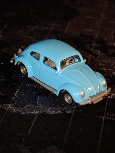 A close-up, eye-level, medium shot captures a blue Volkswagen Beetle toy car positioned on a black surface with a faint world map pattern. The car is facing right, angled slightly towards the viewer. The toy car is detailed, featuring silver bumpers, headlights, and wheels. The interior is visible, showing gray seats and a steering wheel. The background is dark and out of focus, emphasizing the toy car as the primary subject. The location of Dörpen, Germany is not depicted or implied in this image.