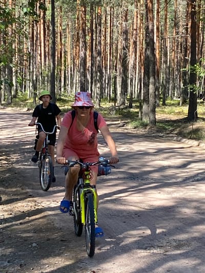In Leipyasuo, Russia, two individuals are cycling on a dirt path through a pine forest. The person in the foreground, on the right, is an adult woman wearing a pink patterned hat, sunglasses, a pink t-shirt, and shorts. She is riding a yellow and black bicycle. To her left and slightly behind is a younger person, likely a child or teenager, wearing a green hat, a black t-shirt, and shorts. This individual is riding a red and white bicycle. Both are on a well-worn dirt path, with tall, slender pine trees lining both sides of the path. The ground is sandy and uneven, with shadows from the trees cast across it. The overall scene depicts a recreational activity in a natural, wooded environment.