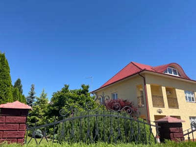 This media file captures a bright, sunny day in Chahor, Ukraine. The scene features a yellow, two-story house with a red tiled roof, set against a clear, vibrant blue sky. The house has several windows and two small balconies with decorative black metal railings. An antenna is visible on the roof.

In the foreground, a black wrought-iron gate with an ornate scroll design is prominent, framing a lush green garden. The gate is supported by brick pillars, one of which is clearly visible on the left side of the image, constructed from dark red bricks. Various types of evergreen trees, including tall cypress-like trees and smaller conical conifers, form a dense backdrop behind the gate and to the left of the house. A deciduous tree with deep red foliage adds a splash of color amidst the greenery.

The overall impression is one of a well-maintained residential property on a pleasant day. The sunlight suggests it is midday or early afternoon. No people or specific activities are depicted. The image appears to be a static shot of the exterior of the house and its surrounding landscape. There is a small sign with text on the house, but it is not legible.