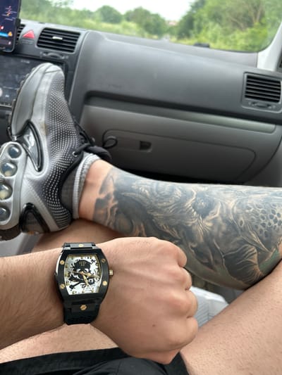 A first-person perspective image shows a person seated inside a vehicle, likely a car, with a tattoo on their left leg and a watch on their left wrist.
The leg, heavily tattooed with a grayscale design depicting a monstrous figure, is positioned in the foreground. A black and gray Nike Air Max Plus sneaker is visible on the foot. The shoe rests on the dashboard, partially obscuring the car's interior features, including an air vent and a touchscreen interface.
The left wrist displays a black and gold skeleton-themed watch with a skull and crossbones motif on the face. The watch band appears to be made of a textured black material.
The vehicle's interior consists of gray plastic components. Outside the car's windshield, green foliage is visible, suggesting a natural environment.
There is no evidence that indicates the location to be Medovo, Bulgaria.