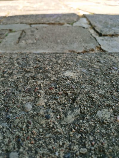 The image is a close-up, low-angle shot of a cracked concrete surface. The foreground consists of coarse gravel and sand embedded in concrete, with small pebbles of varying colors. In the background, larger, lighter-colored concrete slabs are visible, with significant cracks and some sparse green vegetation growing in the crevices. The lighting suggests it is daytime, with shadows falling across the scene. The overall impression is of an aged and weathered outdoor paved area, possibly a sidewalk or patio. No people or specific landmarks of Švenčionys, Lithuania are identifiable.