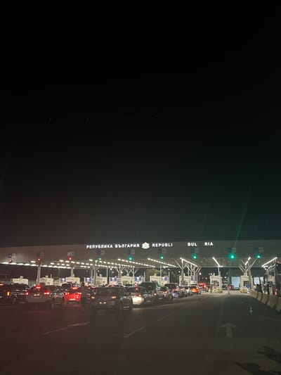 At nighttime, the Kapitan Andreevo border crossing in Bulgaria is depicted with multiple lanes of traffic backed up, waiting to pass through. The border crossing infrastructure is illuminated, with prominent signs reading "РЕПУБЛИКА БЪЛГАРИЯ" and "REPUBLIC OF BULGARIA". Numerous vehicles, primarily passenger cars and some larger vehicles, are stationary in their respective lanes. Traffic lights are visible, with some showing red and others green, indicating the ongoing processing of vehicles. Several individuals, likely border officials or travelers, are observable near the booths and in the vicinity of the vehicles. The scene suggests a busy border crossing with a significant queue of vehicles.
