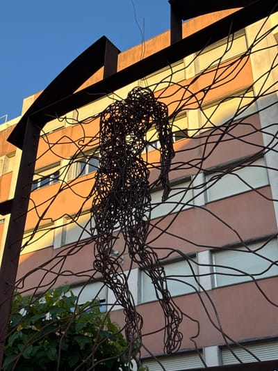 The image is a low-angle shot taken in Vinaròs, Spain. The primary subject is a hanging sculpture made of twisted dark metal wire, resembling a human figure. The sculpture hangs from a dark metal structure which forms a partial frame on the lower left.

Behind the sculpture is a multi-story building with alternating salmon and white panels on its façade. Windows are visible on the building.

To the right of the sculpture are green leafy plants. The sky is blue in the lower left. The scene is lit by sunlight.