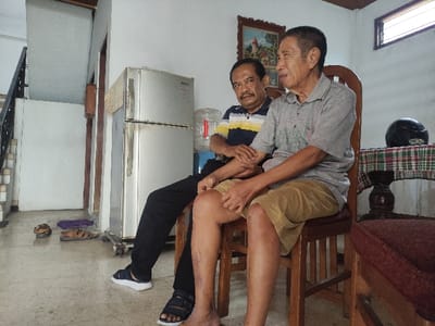 This indoor photograph, taken in Lamongan, Indonesia, depicts two men seated in what appears to be a living room or common area of a home.
On the left, a man with dark hair and a mustache is seated on a wooden chair. He wears a polo shirt with yellow, white, and blue horizontal stripes, dark pants, and dark sandals. His hands are clasped, and he is looking towards the right side of the frame. Beside him, to the right, sits an older man with short, graying hair. He is wearing a light gray polo shirt and light brown shorts, and is barefoot. He also looks towards the right. Both men appear calm and relaxed.
The background features household items and architectural elements. A silver-colored, two-door refrigerator stands prominently behind the men. Next to the refrigerator is a water dispenser with a large, clear water bottle on top. To the far right, a small wooden table covered with a plaid tablecloth (featuring green, red, and white checks) holds a black motorcycle helmet. A high window above the table allows natural light into the room, indicating it is daytime.
On the left side of the image, a staircase with a dark railing ascends. Several pairs of sandals or slippers are neatly placed on the light-colored tiled floor near the base of the stairs. Wooden doorframes or posts are visible, and the walls are painted white. A framed picture, depicting a landscape or building, hangs on the wall above the men's heads. There is no visible text in the image.