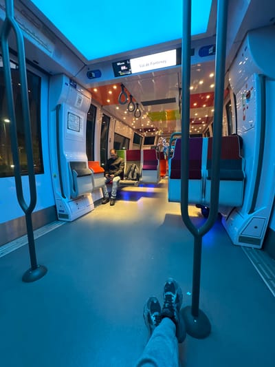 The image, taken in a vertical orientation but depicting a horizontally oriented scene, captures the interior of a modern train or tram carriage, likely in the evening or at night. The perspective is from a standing passenger, whose feet, clad in dark sneakers and gray sweatpants, are visible in the foreground. A dark metal handrail stretches across the lower-mid section of the frame.
The carriage is brightly illuminated by a mix of cool blue and warm yellow lights. A large, rectangular blue light panel dominates the left side (which would be the ceiling if the image were rotated), casting a cool glow. Numerous small, warm-toned lights are embedded in the ceiling and along the walls, creating a scattered, reflective pattern. Blue accent lighting highlights the floor level along the seating structures.
Further down the spacious, blue-gray floored aisle, another passenger is seated on the left, appearing to be looking down at a device. The seats are modern, with segments of red, purple, and gray upholstery. White overhead panels and grab handles are visible throughout the carriage.
A prominent display screen on the left side of the carriage clearly shows the text "Val de Fontenay," likely indicating the next stop or destination. Below this, smaller, illegible text is present. A small map or informational graphic is affixed to a wall panel near the seated passenger. The interior is clean, modern, and features a sleek design. The dark windows suggest it is nighttime outside, enhancing the interior lighting's effect.