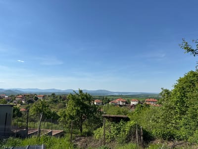 The image depicts a sunny, rural landscape in Bokilovtsi, Bulgaria. A view of the village is visible, with houses clustered among trees and vegetation. The houses feature orange-red tiled roofs, characteristic of the region. In the distance, a body of water is discernible, bordered by rolling hills and mountains. The foreground includes a mix of greenery, including trees, bushes, and long grass, some of which appears unkempt or wild. A section of chain-link fence and concrete steps are visible on the left side of the image, likely indicating the boundary of a private property. The sky is a clear, bright blue, with only a few wisps of clouds visible.