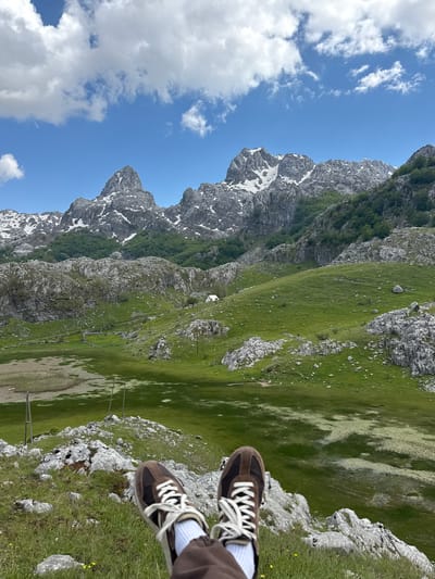 The image depicts a mountain landscape in Brskut, Montenegro. Jagged, rocky peaks with patches of snow are visible under a blue sky with scattered clouds. A green, grassy hillside slopes down towards a valley. In the foreground, a pair of brown shoes with white laces are visible, as if someone is sitting at the edge of a rocky outcrop overlooking the landscape. Power lines stretch across the valley floor.