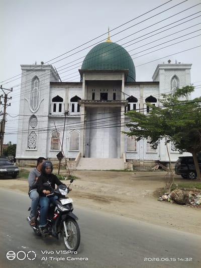 A wide shot captures a mosque with a large green dome, accented by white ornate minarets, in Keudelapang, Indonesia. The mosque appears to be under construction, with scaffolding visible on its facade. In the foreground, two individuals ride a black motorcycle along a paved road. The rider wears a helmet, and the passenger is seated behind him. A black car is parked on the left side of the road, and a black SUV is visible on the right. The ground is dusty, with scattered debris and a large woven basket filled with trash to the right of the mosque. Overhead, a network of power lines crisscrosses the frame, indicating an urban or semi-urban setting. The sky is overcast. The lower right corner of the image displays a timestamp: "2026.06.15 14:32".