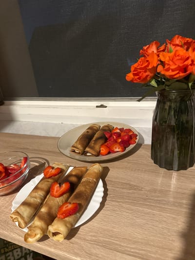 This image captures a static indoor scene in Arzberg, Germany, displaying prepared food and floral decoration on a wooden countertop positioned before a window. The countertop is a light brown, laminated surface exhibiting a distinct horizontal wood grain.
In the background, a white window sill and frame are visible. Behind this, a dark, fine-mesh screen covers the window opening, suggesting either night-time or an obscured outdoor view.
On the left side of the countertop, a clear glass bowl contains a quantity of diced red strawberries. Adjacent to this, in the central foreground, a white plate with a subtly scalloped edge holds three rolled crepes or pancakes. These crepes are golden-brown and appear filled; two are topped with a half-strawberry precisely cut into a heart shape, and the third with a plain half-strawberry. Further back on the countertop, a light-colored, irregularly shaped plate presents three additional rolled crepes alongside a pile of fresh, diced red strawberries.
To the right of the scene, a dark, translucent glass vase with vertical fluting contains a bouquet of vibrant orange roses. The roses are in various stages of bloom, with discernible green stems and foliage.
Artificial indoor lighting illuminates the scene, casting soft shadows across the items. Partially visible Cyrillic text, including characters resembling "ЬДЕЩ" and "ЗВАК", is present on an object located beneath the left edge of the countertop. No individuals are depicted in the image.