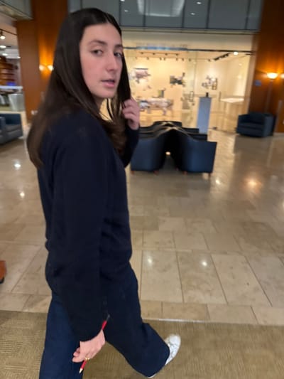 The image captures the interior of a building identified as the Rose Art Museum at Brandeis University in Waltham, Massachusetts. A female individual is positioned in the foreground, mid-stride, moving from left to right while her upper torso and head are turned towards the camera. She has long, dark hair parted centrally. She is dressed in a dark blue long-sleeved top and dark blue pants, with white sneakers on her feet. Her right hand is raised to her hair near her temple. Her left hand holds a red and white pencil, and a gold-colored ring is visible on her left ring finger. The flooring consists of light-colored, possibly beige, square tiles, which reflect ambient lighting. A textured, light brown rug is partially visible in the lower left foreground. In the background, several dark, curved modular seating units are arranged on the tiled floor. Behind these units, a glass-enclosed exhibition space is visible, identified by the sign "Rose Art Gallery" above its entrance. Abstract artworks are displayed inside the gallery on white walls. A dark blue armchair and a standing lamp with a warm glow are also present in the background.