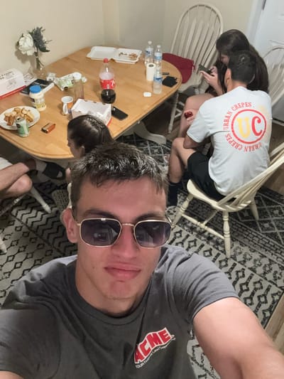 The image depicts a group of people gathered around a dining table in what appears to be a casual indoor setting, likely a home or vacation rental in Ocean City, United States of America. The scene is framed as a selfie taken by a young man in the foreground wearing sunglasses and a gray t-shirt with a red and white logo that reads "ACME". He is looking directly at the camera. Behind him, the dining table is laden with food containers, bottles of soda and water, a bouquet of flowers in a vase, and other assorted items. Several other individuals are visible, including two women and at least one other person whose legs are partially in view. One woman is seated, looking at her phone, while another is partially obscured. The lighting suggests it is either evening or an indoor space with artificial light. The overall atmosphere appears relaxed and informal.