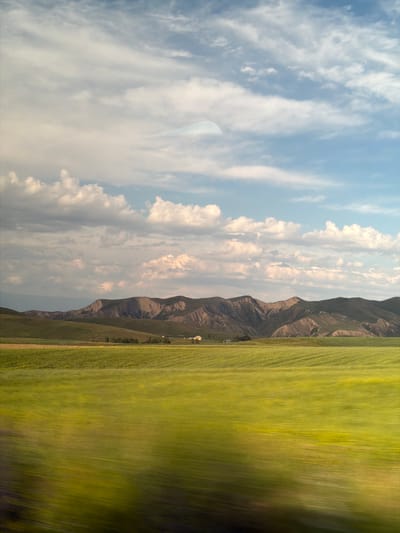 This is a vertical landscape photograph captured in Toguz-Bulak, Kyrgyzstan. The image depicts a vast, green field in the foreground, exhibiting motion blur that suggests movement, possibly from a vehicle. In the mid-ground, a range of rugged, brown mountains rises majestically, their slopes textured and contoured. A few small, white buildings are visible nestled at the base of the mountains, hinting at a rural settlement. The sky above is a mix of blue and white, with scattered clouds, some of which are illuminated by sunlight, indicating it is likely daytime. The overall mood is one of natural beauty and expanse, with the motion blur adding a dynamic element to the serene landscape. There is no visible text in the image.