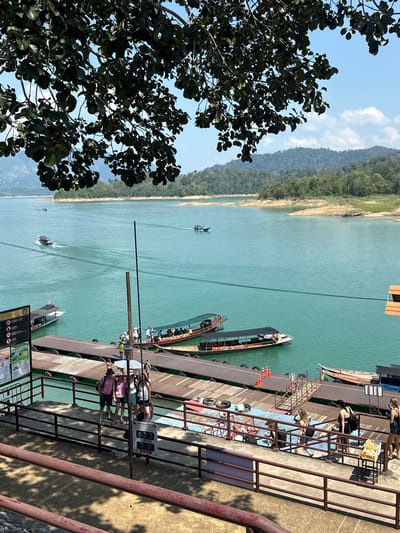 The image, taken from an elevated vantage point, captures a waterscape at the city of Ban Ta Khun, Thailand. The scene encompasses a body of water, appearing to be a lake or reservoir, of a light turquoise hue. Boats are visible on the water, including longtail boats docked at a series of piers, indicative of tourist activity.
People are present on the piers, some seated and others standing. The piers themselves are constructed of wood or similar material, showing signs of wear. A metal railing runs along the side of the elevated platform, offering a safety barrier and contributing to the man-made elements of the landscape.
The shoreline features lush, green vegetation, extending into a forested mountain range in the background, where the sky is visible with white, puffy clouds. The sky is blue above. A large tree with dense, dark green foliage dominates the left foreground of the image, partially obscuring the view.