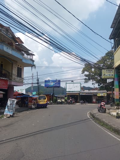 The image shows a street scene in an unidentified location, likely in Southeast Asia, given the architecture and signage. The road is asphalt with visible wear. A yellow and purple van is parked on the left side of the road. Numerous motorcycles are parked along the roadside. Various small shops and food stalls line the street, with several signs in Indonesian. Overhead, there's a dense network of electrical wires. In the background, a hill is visible, with an advertisement billboard erected on its slope. The sky is partly cloudy.