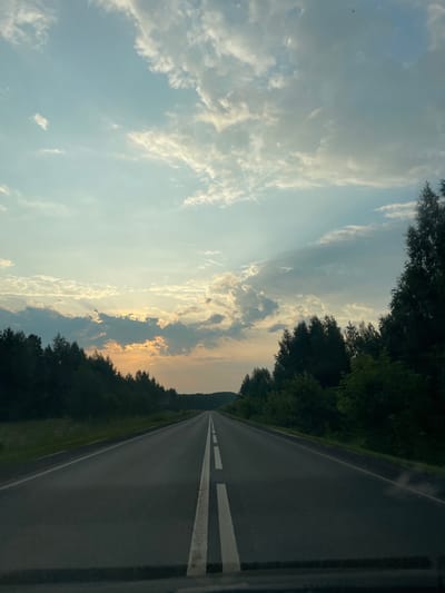 A straight asphalt road with white double yellow lines runs towards the horizon, bordered by dark green trees on both sides. The sky above is a pale blue with scattered white and grey clouds, and the sun is setting or rising, casting a warm glow through the clouds on the left side of the frame. The overall scene depicts a rural or semi-rural road in Novoe Mart'yanovo, Russia, during twilight.