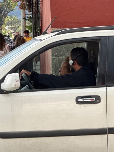 Inside a white vehicle, a man with headphones is focused on driving, his hands on the steering wheel. Beside him, a lighter-haired person looks down. The scene is framed by the open car door, revealing a glimpse of an exterior setting with people, foliage, and buildings. A red wall is seen behind the car. The door has a vertical handle.