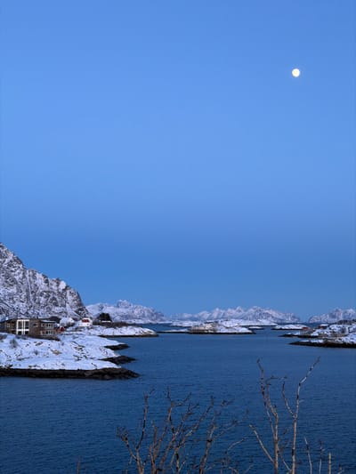 The photo presents a landscape with a clear blue sky where a round moon is visible. The lower half reveals snow-capped mountains and small buildings nestled near a body of water. The water appears dark blue, possibly a fjord or lake. Sparse branches of a tree are noticeable on the right side of the image. The scene evokes a sense of coldness and tranquility.
