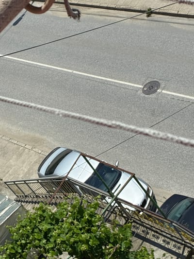 This is an overhead, tilted, high-angle shot of a street scene in Vlahovo, Bulgaria. A light-colored car is parked at an angle on the asphalt road, which has a white lane marking. A metal fence, likely a balcony or staircase railing, is visible in the foreground, partially obscuring a cluster of green leafy plants. To the right of the light-colored car, the rear of a dark-colored car is visible. In the background, a concrete sidewalk runs alongside the road, and a small plant sprouts from a crack in the pavement. A circular metal manhole cover is present on the road. Two taut, thin wires are stretched across the frame, also partially obscuring the view. There are no people clearly visible in the frame.