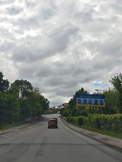 A street view in Nemyriv, Ukraine, under a cloudy sky. The road leads uphill with an orange car traveling away from the camera. A billboard stands on the right side of the road, featuring portraits with Ukrainian text above. Vegetation lines both sides of the road, with trees on the left and bushes on the right. A few buildings are visible in the distance.