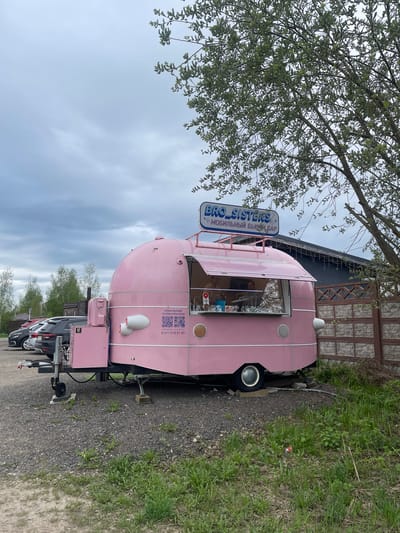 The image shows a pink "BRO SISTERS" mobile beauty salon in the city of Хлябово, Russia. The salon is a small trailer parked on a gravel path next to a patch of grass. A sign with the salon's name, "BRO SISTERS," is mounted on top. A tree with light green foliage is visible in the upper left corner of the image. Cars are parked behind the trailer. A building is behind the trailer, with a stone-clad facade. The sky is overcast.