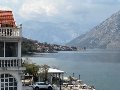This image depicts an exterior scene in the city of Bogdašići, Montenegro, showcasing a bay and surrounding mountains under an overcast sky. In the foreground on the left, a multi-story white building with a red-tiled roof and a prominent white balustraded balcony is visible. Below, a white Citroen C4 Cactus SUV is parked on a paved area. Adjacent to the parked vehicle, an outdoor dining establishment extends towards the water, featuring multiple white gazebos, tables, and chairs, some covered. Several black street lamps with white tops are interspersed among the dining sets. The bay occupies the central and lower right portions of the frame, appearing calm with a ladder descending into the water from a concrete pier. Along the distant left shoreline, a small settlement of buildings with red-tiled roofs is situated at the base of a tree-covered hill. Steep, rocky mountains rise directly from the water on the right side of the image, receding into the background. Several small boats are moored in the water near the distant shore. No individuals are visible in the scene.