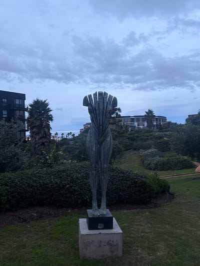 A landscape photograph captures a statue of a stylized human figure, possibly made of metal, with a head composed of vertically oriented, flat elements. The statue stands on a concrete pedestal in a garden setting with green grass and diverse vegetation, including bushes and trees. In the background, buildings are visible against a cloudy sky. The presence of palm trees suggests a coastal environment. Given the stated location of Milnerton, South Africa, the scene is likely a public park or garden featuring the statue. There are no people visible.