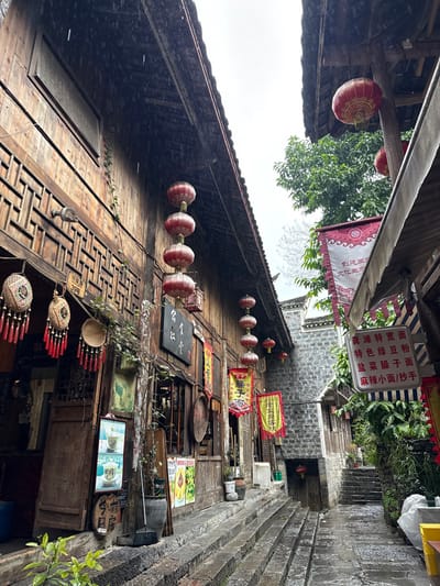 The image depicts a narrow, wet stone-stepped alleyway in Gongtan, China, under an overcast sky. The pathway ascends gradually into the distance, flanked by traditional multi-story buildings.

On the left, a building constructed of dark wooden planks features intricate carved lattice work and an overhanging dark tiled roof. A vertical string of five round red Chinese lanterns hangs from the eaves, with additional lanterns visible further along the facade. Decorative woven items with red tassels are suspended near a doorway, which also has a small white sign marked "39号" (Number 39). A black plaque with white Chinese characters and multiple yellow and red banners displaying Chinese text are affixed to the building's exterior. A brown broom leans against the wall, and potted plants sit at the base.

On the right, another building with a similar overhanging tiled roof and wooden elements is partially obscured by dense green foliage. A white sign with blue text and Chinese characters, advertising food items such as "特色绿豆粉" (Special Mung Bean Noodles) and "盐菜臊子面" (Salted Vegetable Minced Pork Noodles), hangs from an awning. Further into the alley, a building section with grey stone block construction is visible, adorned with additional red lanterns and banners. The stone steps and ground surfaces appear wet, indicating recent precipitation. No individuals are present in the scene.
