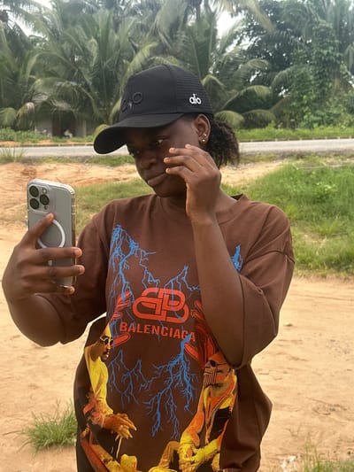 A young Black woman, wearing a black trucker hat and a brown t-shirt with a graphic print, stands outdoors and holds a smartphone in her left hand, taking a selfie. The woman has her right hand raised to her face, touching her eye. She is looking at the phone screen, which is not visible. The t-shirt features a large graphic with blue lightning bolts and text that reads "BALENCIAGA". The woman is positioned in the foreground against a backdrop of palm trees and greenery. The ground appears to be sandy and dirt. The location is identified as Ganyingbo, Nigeria.