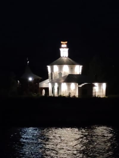 A night-time, slightly blurred, eye-level shot depicts a brightly lit, white, multi-tiered church structure situated in Uglich, Russia, likely near a body of water, as evidenced by the rippling, moonlit reflections in the foreground. The church is the central focus, its windows glowing with warm light against the dark sky. To the left of the main structure, a darker, conical-roofed building, possibly a bell tower or another church building, is partially visible. The main church features multiple levels with arched windows, and its topmost dome is illuminated, crowned with a golden finial. The scene is devoid of any people or discernible activity, creating a serene and somewhat isolated atmosphere.