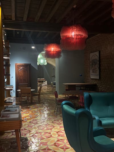 The image presents an unoccupied interior space, identified as a lounge or communal area, located in Arles, France. The setting is illuminated primarily by artificial light from multiple sources. The ceiling features dark, exposed wooden beams. The floor is tiled with a complex geometric pattern utilizing shades of yellow, orange, red, and light pink.

Three prominent, large pendant light fixtures are suspended from the ceiling: two in the mid-ground appear red and have a textured, undulating design, while a third, smaller fixture visible through an archway in the background, emits a soft green light. The walls exhibit a dual composition: sections are painted a light blue-gray, while others feature exposed light brown brickwork.

Furniture within the immediate view includes a teal velvet upholstered armchair positioned with its back to the viewer in the foreground, adjacent to a matching two-seater sofa. To the left, a small circular wooden table is accompanied by two light-toned wooden chairs. A tall, narrow wooden bookshelf, stocked with various publications, stands against the left wall. Nearby, a tall, ornate dark wooden door is visible. Further into the room, a rectangular wooden table is set with several chairs upholstered in red fabric. On the right, a framed black-and-white poster hangs on the exposed brick wall. An arched passage in the background leads to an additional, dimly lit area containing more seating and tables. No individuals are present in the scene.
