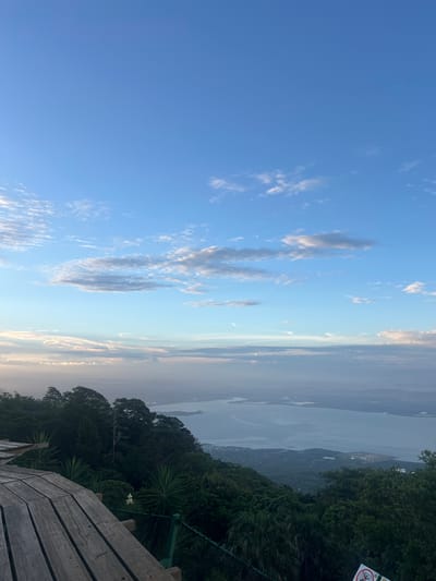 This is a wide-angle, eye-level shot of a scenic overlook in El Cacao, El Salvador, taken from a wooden platform. The foreground features the weathered wooden planks of the platform, some showing signs of wear. Beyond the platform, dense green foliage and trees dominate the left side of the frame. A white flower with green leaves is visible amidst the greenery. A green fence runs horizontally across the middle ground.

The background reveals a vast expanse of water, likely a lake or bay, with several islands or landmasses dotting its surface. The water is calm and reflects the soft light. Distant mountains or hills are visible on the horizon, shrouded in a hazy, atmospheric perspective. The sky above is a gradient of blue, transitioning from a clear, deep blue at the top to lighter shades with scattered white clouds near the horizon. The lighting suggests it is either early morning or late afternoon, with a gentle, diffused light. The scene conveys a sense of tranquility and natural beauty.