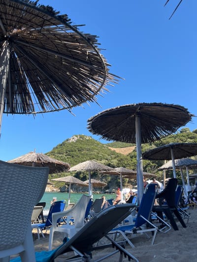 The image depicts a beach scene with numerous straw umbrellas and lounge chairs set up on the sand. The foreground is dominated by the textured surfaces of a white plastic chair and a reclined lounge chair with a blue towel. Behind these, a row of lounge chairs, some blue, some black, and some white, are arranged under various straw umbrellas. The sea is visible in the background, with calm, turquoise water lapping at the shore. Several people can be seen on the beach and in the water, some relaxing on chairs, others wading. A lush, green, tree-covered hill rises behind the beach, creating a scenic backdrop. The sky is a clear, bright blue. The overall impression is one of a tranquil summer day at a coastal resort. The scene is likely located in Νέα Ηρακλείτσα, Greece, a region known for its beautiful beaches and clear waters.