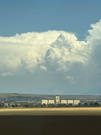 In Svetlograd, Russia, a vast agricultural landscape unfolds under a dramatic sky. The foreground is dominated by two distinct fields: one a dark, rich earth, the other a sun-drenched yellow, likely crops. Beyond these fields, a town stretches across a gentle hillside, dotted with buildings and trees. Prominently featured in the middle ground are several large, modern industrial structures, possibly grain elevators, with one tall tower standing out. These structures are set against the backdrop of a hazy horizon and rolling hills. Above, an immense, dynamic cloud formation fills the upper portion of the image, its white, cumulus shapes contrasting sharply with the clear blue sky peeking through. The overall scene suggests a rural or semi-industrial setting in a vast, open expanse under a powerful sky.