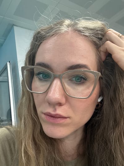 A woman with long, wavy blonde hair is looking at the camera. She is wearing glasses with a light-colored frame. Her right hand is resting on her head. She has green eyes and pink lips. A white wireless earbud is visible in her right ear. The background shows a blue-and-white tiled wall and a partially visible silver object, possibly a window frame. The scene is not identifiable to be in the city of Gungoren, Turkey.