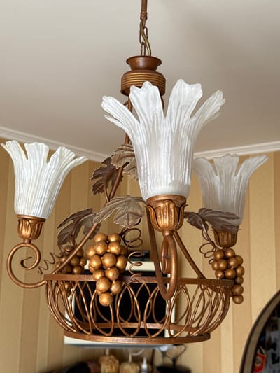 A close-up, low-angle shot captures an ornate, vintage-style chandelier hanging from a textured ceiling. The chandelier is made of a coppery-bronze metal and features three tulip-shaped frosted glass shades, each with delicate vertical ridges. Decorative elements include stylized grapevine leaves and clusters of faux golden grapes adorning the fixture. The background consists of a wall with vertical yellow stripes.